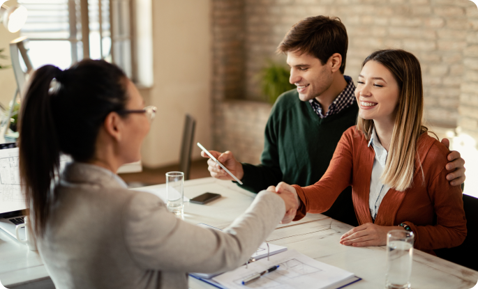 A professional woman in a grey blazer shakes hands with a young woman across a desk. A young man sits next to the woman, smiling as he holds a digital tablet. They are in a modern, sunlit office with brick walls, appearing to finalize a deal or agreement.