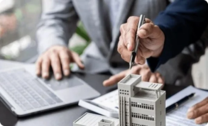 Two professionals collaborate over a physical architectural model and a laptop at a desk. One person points a pen toward the miniature building while the other works on a computer, symbolizing real estate development or design planning.
