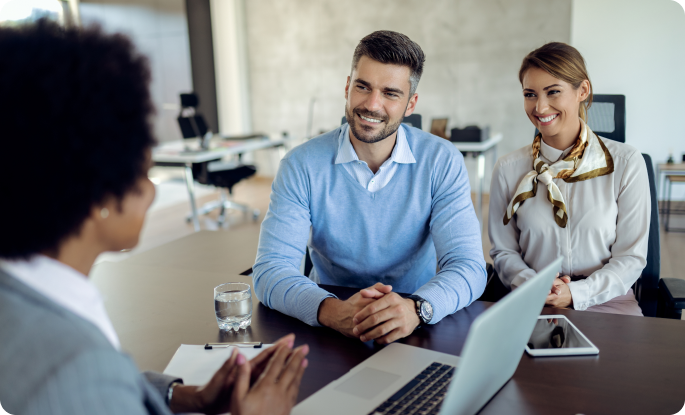 A professional woman in a grey suit meets with a smiling young couple across a dark conference table. The group is in a bright, modern office with a laptop and digital tablet open for their discussion