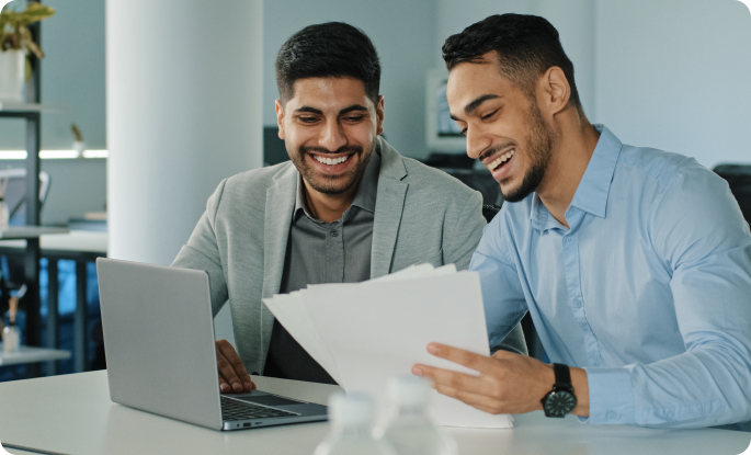 Two smiling colleagues reviewing paperwork together in a modern office setting with a laptop.