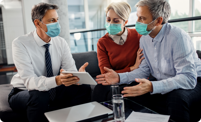 Three people wearing light blue surgical masks sit around a glass coffee table. A man in a white shirt and tie holds a tablet while a couple listens intently.