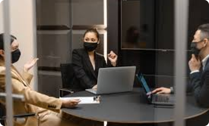 Three people wearing black face masks sitting around a dark circular table in a minimalist office setting, engaged in a discussion with laptops present.
