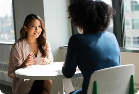 Two women engaged in a professional meeting or interview at a small round white table near a window. The woman on the left is speaking and gesturing with her hands.